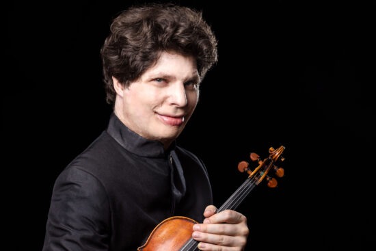 violinist Augustin Hadelich, wearing all black, holds his violin in rest position against his chest.