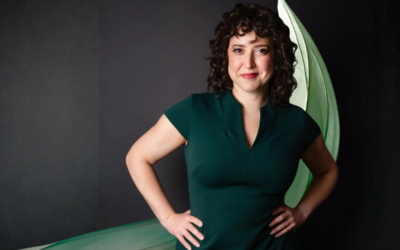 Amy Maude is a white woman with dark, curly hair. She stands here facing the camera straight on with her hands on her hips and a slightly sly smile. She is wearing a dark green dress which is enhanced by an artistic green swoosh of fabric falling behind her.