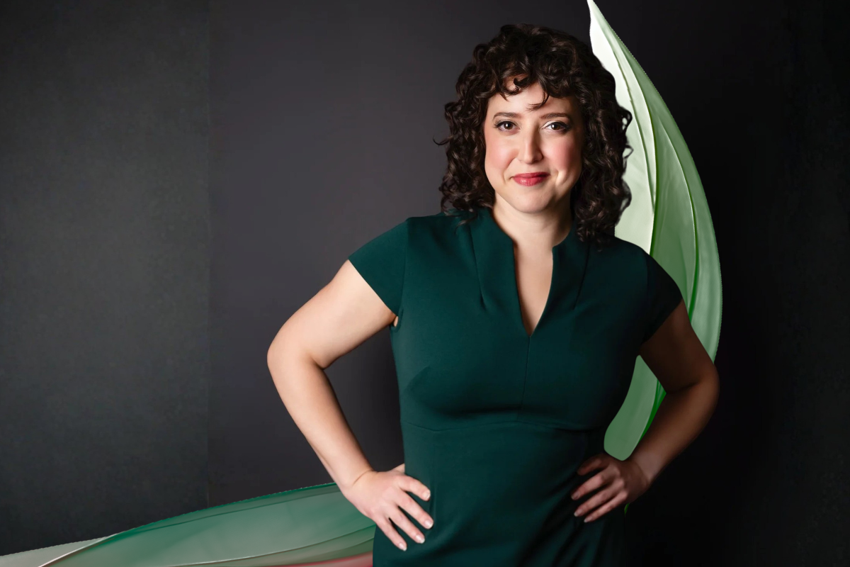 Amy Maude is a white woman with dark, curly hair. She stands here facing the camera straight on with her hands on her hips and a slightly sly smile. She is wearing a dark green dress which is enhanced by an artistic green swoosh of fabric falling behind her.