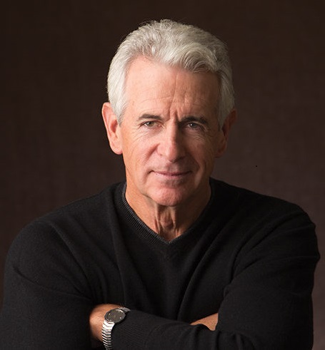 Actor James Naughton sits in front of a dark backdrop with his arms crossed. 