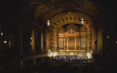The NHSO plays on a warmly lit stage at Woolsey Hall. The hall is shrouded in black and the golden pipes of the organ rise up dramatically above the orchestra onstage.