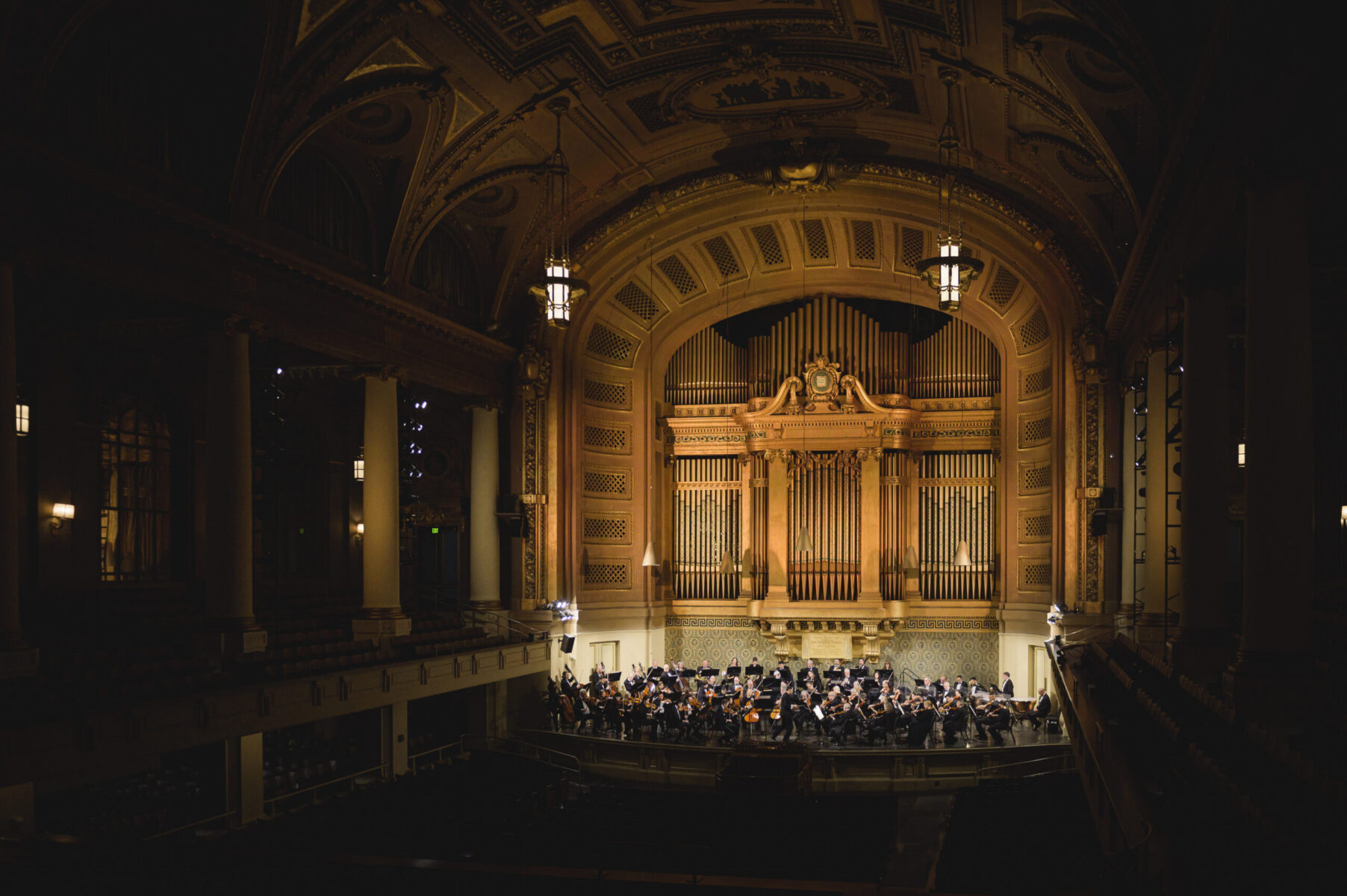 The NHSO plays on a warmly lit stage at Woolsey Hall. The hall is shrouded in black and the golden pipes of the organ rise up dramatically above the orchestra onstage.
